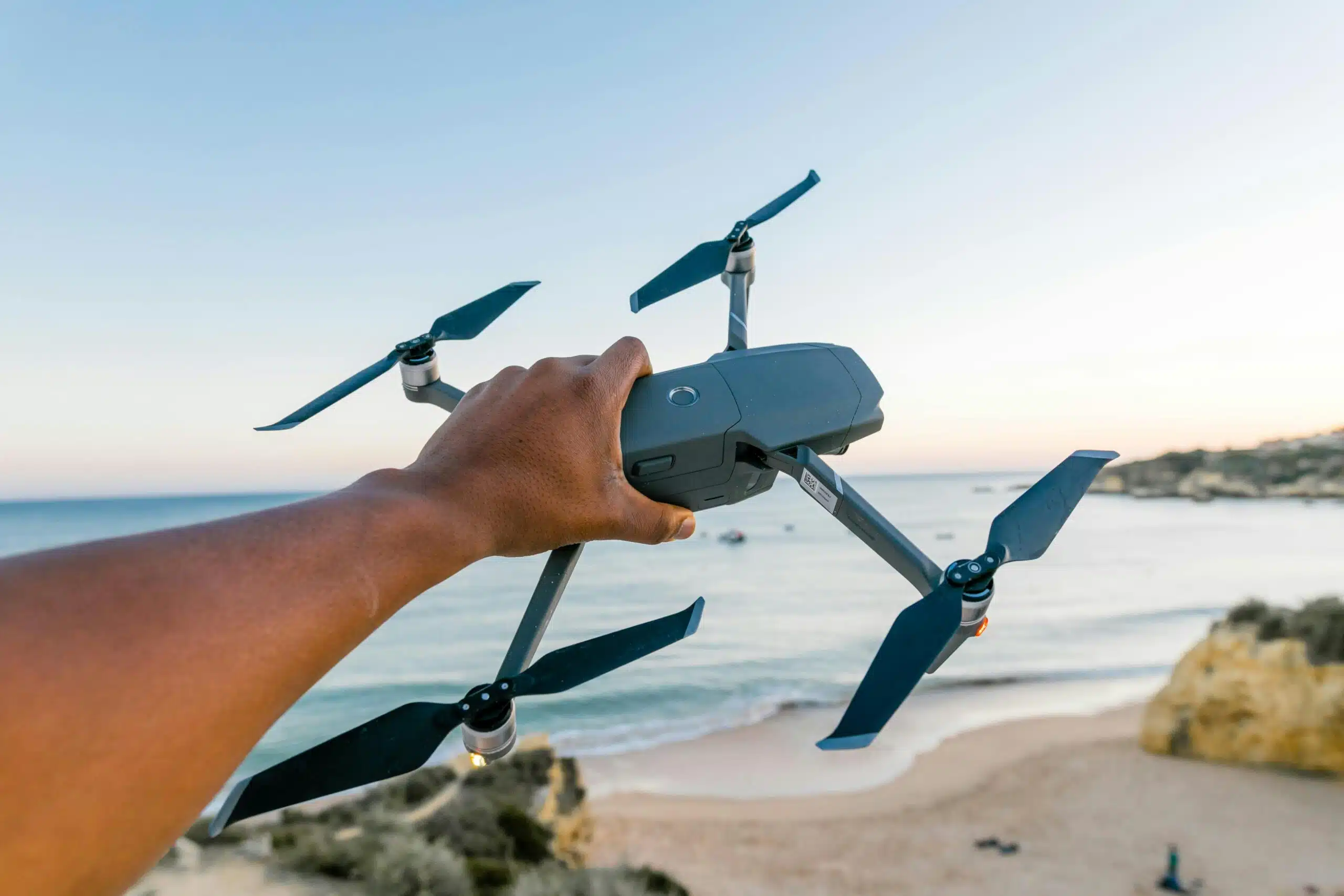 Sky Scan Surveys | A Guide to DJI Assistant 2: Unlocking the Full Potential of Your Drone A hand holding a drone over a beach in Albufeira, Portugal, during sunset, showcasing technology and travel.